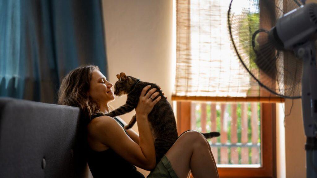 Young woman with a gray cat in her arms sitting at home on the floor in front of a fan, escape from the summer heat concept